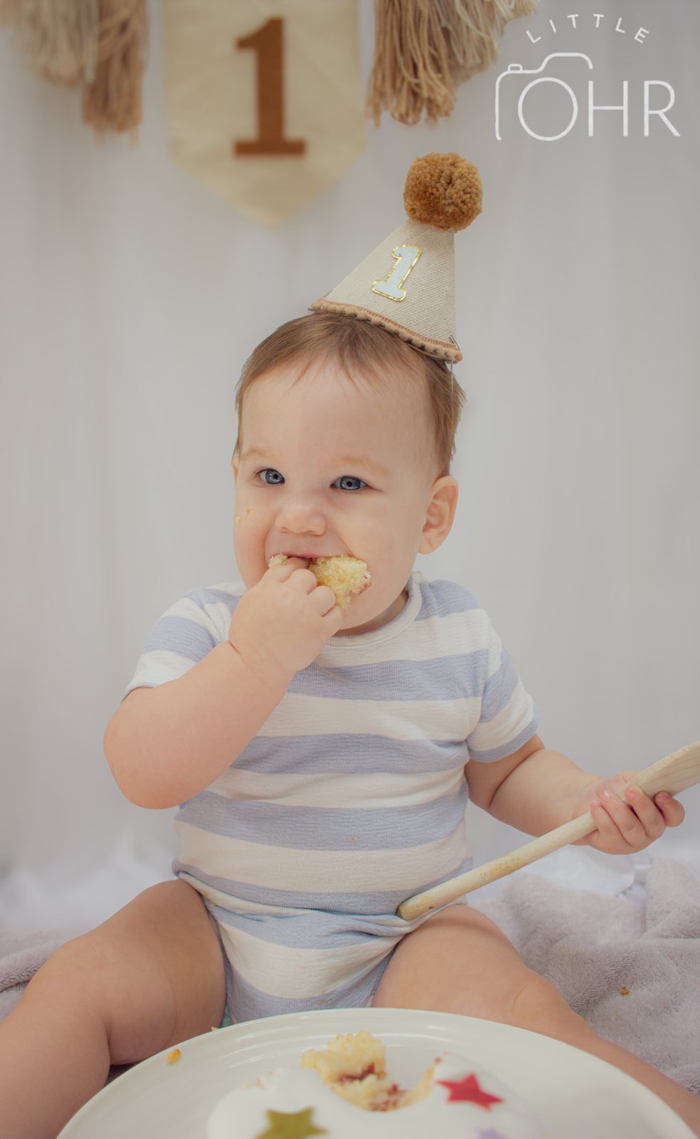 Baby in a striped romper and party hat eating cake, holding a wooden spoon