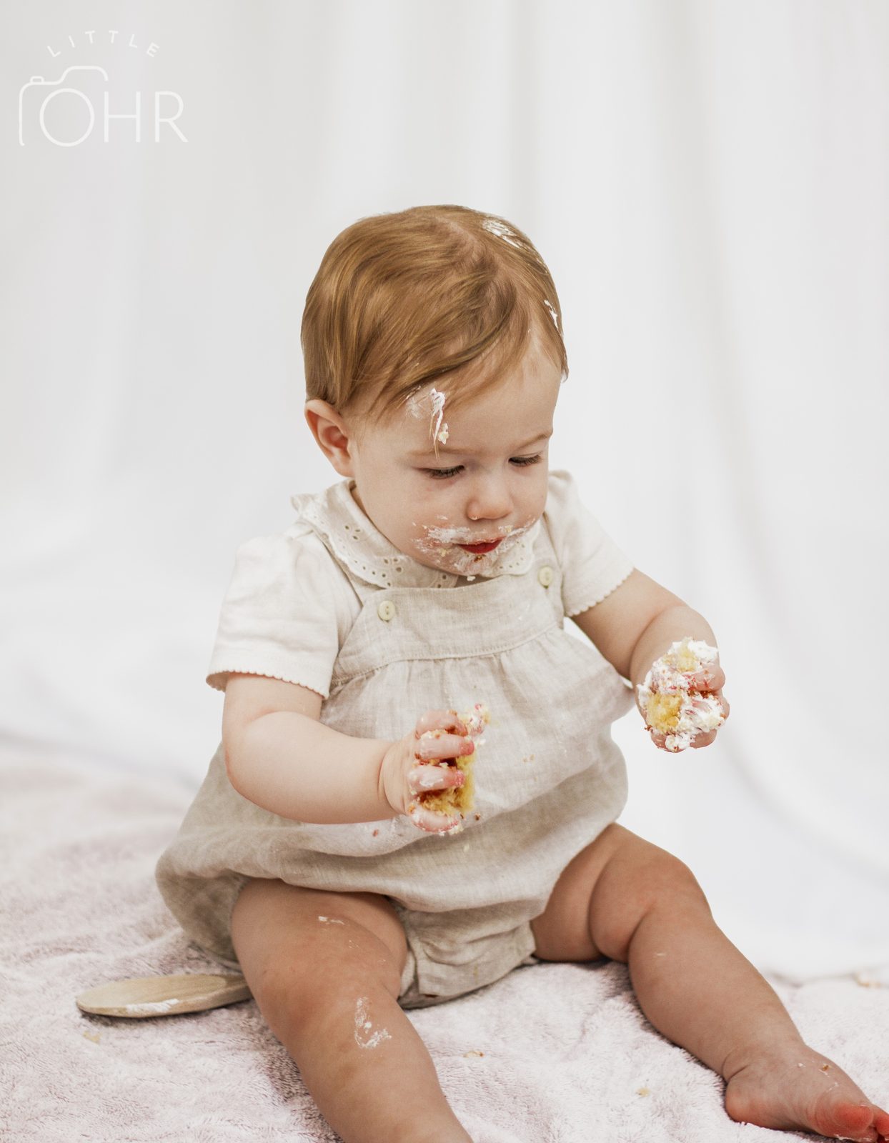 Baby in a linen romper at a cake smash, cake in both hands and on their face