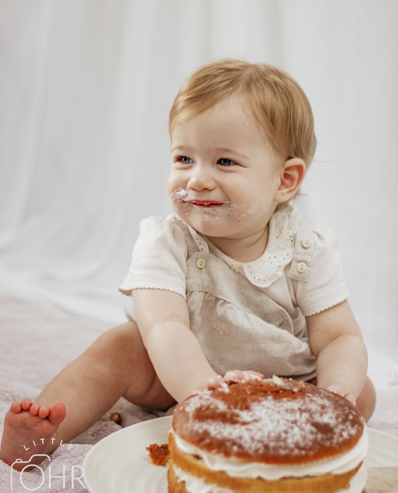 Baby grinning with cream on their face, hands on a sponge cake