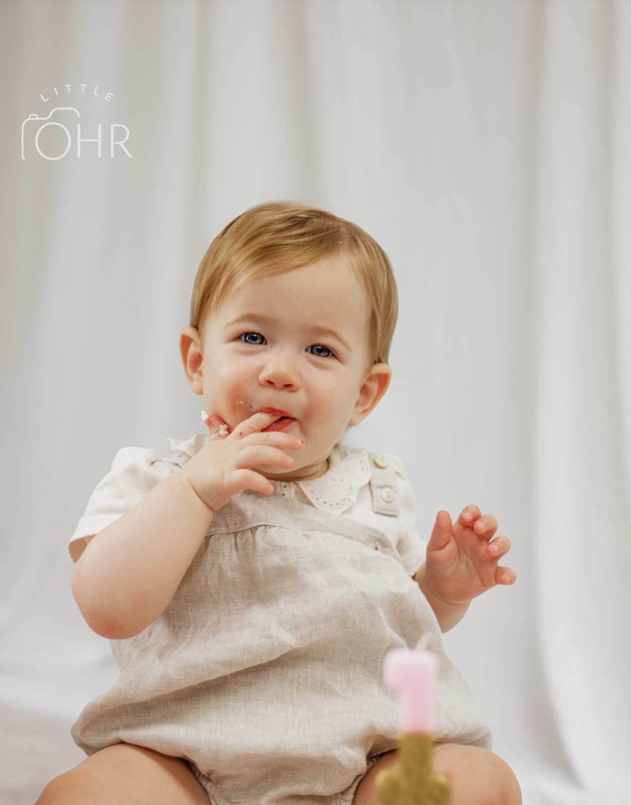 Baby in a linen romper licking their fingers beside a number one candle