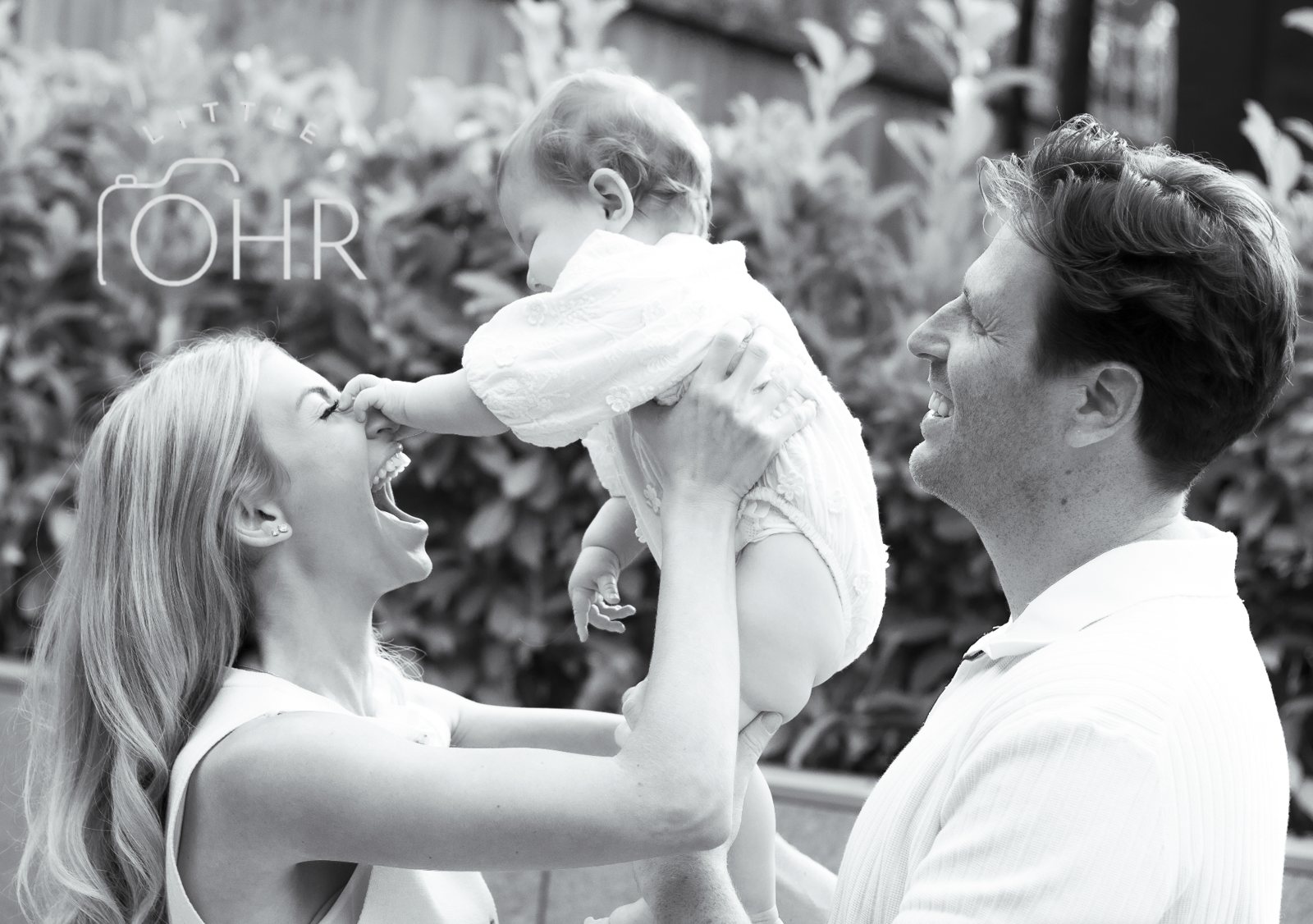 Black and white photo of mum and dad lifting their baby between them in the garden