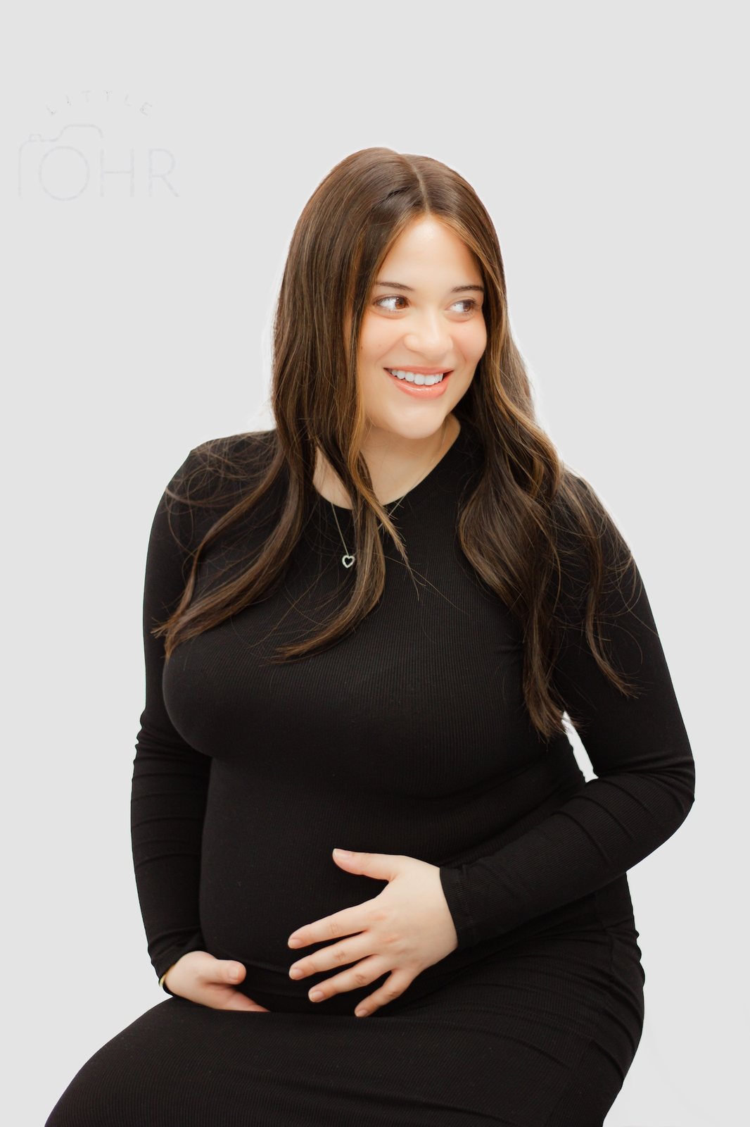 Expectant mum sitting in a black dress with her hands on her bump, smiling to the side