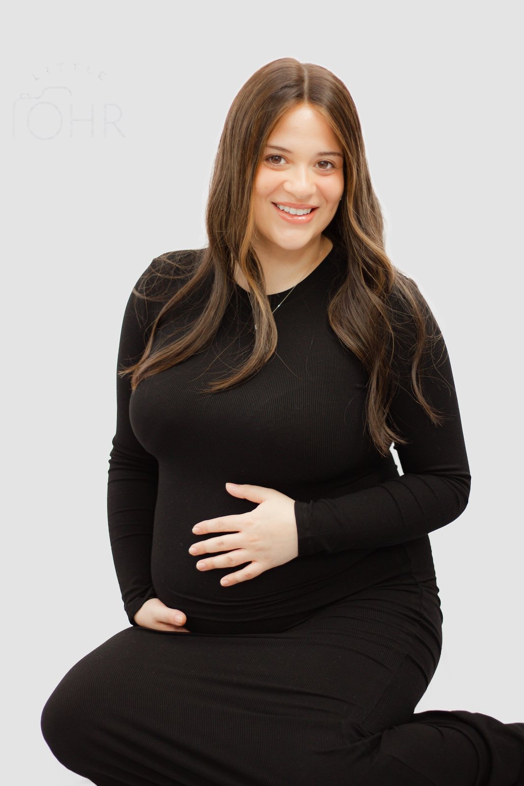 Expectant mum kneeling in a black dress, smiling at the camera with her hands on her bump