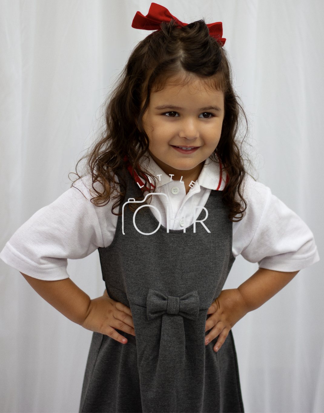 Little girl with a red bow, hands on hips, in a grey school pinafore