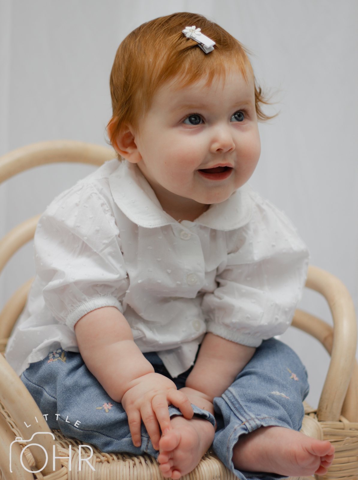 Red-haired baby girl with a bow in her hair, sitting in a little cane chair and smiling