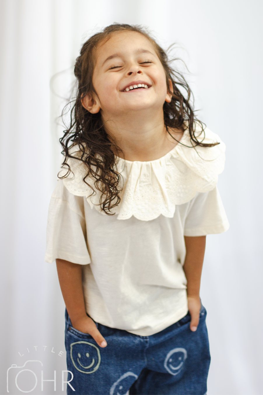 Young girl in a cream top and blue jeans, hands in her pockets, grinning
